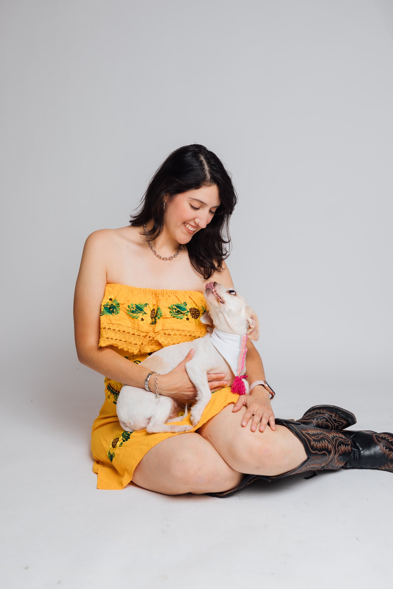 A latin woman sitting on a white studio floor wearing a traditional Mexican yellow strapless dress with green floral embroidery and holding a small white dog wearing a traditional Mexican bandana from Firulais Co. She is also wearing black cowboy boots against a plain light gray background.